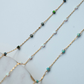 Gold chain necklace with green and blue gemstones on a light blue background