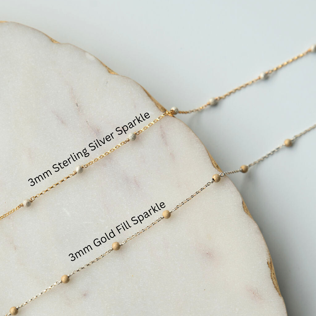 Two gold necklaces with '3mm Sterling Silver Sparkle' and '3mm Gold Fill Sparkle' labels on a marble surface.