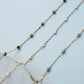 Gold chain necklace with green and blue gemstones on a light blue background