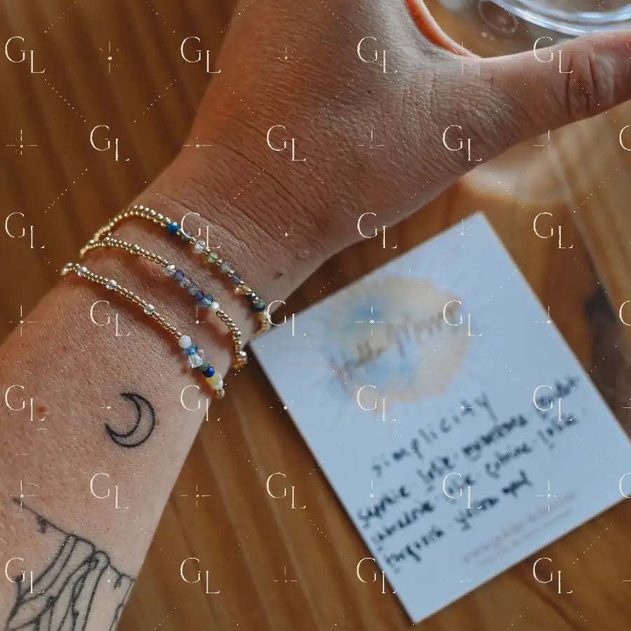 Hand wearing multiple gold bracelets with a card and glass of tea in the background