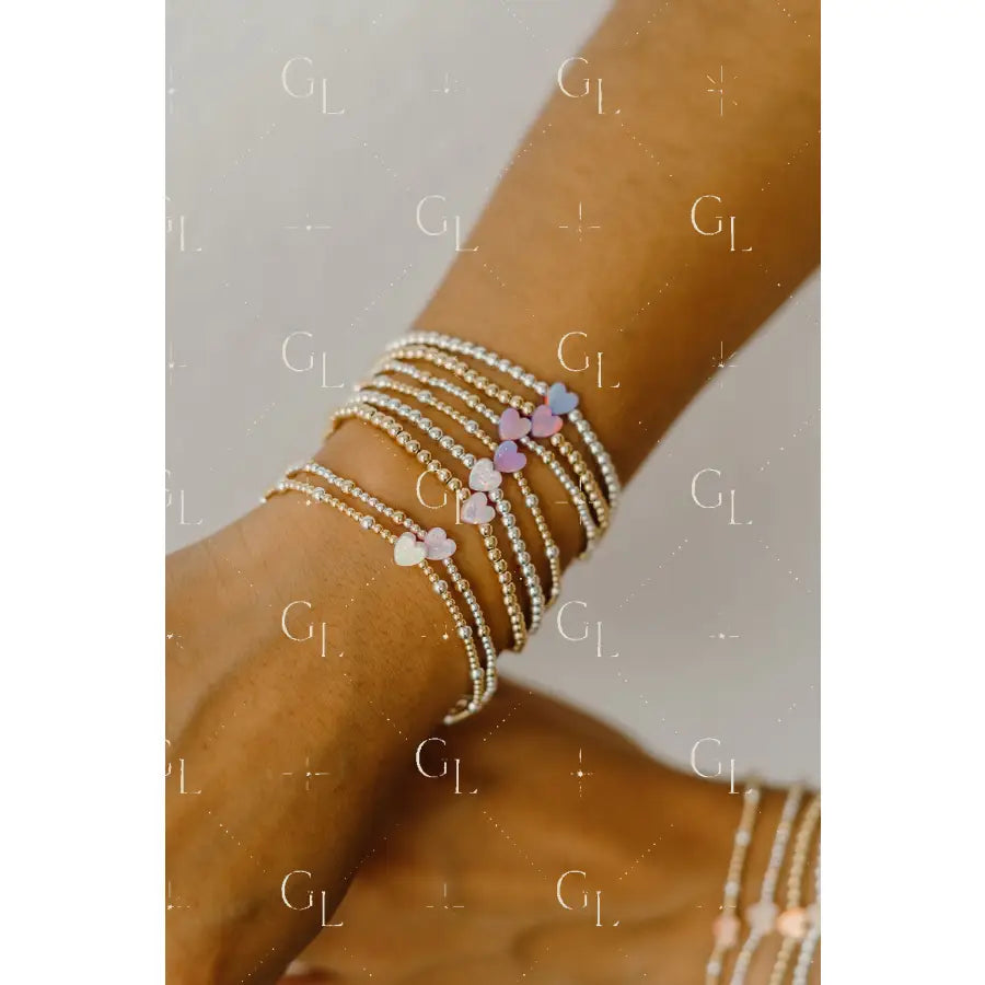 Close-up of a wrist wearing multiple beaded bracelets with heart-shaped charms on a neutral background.