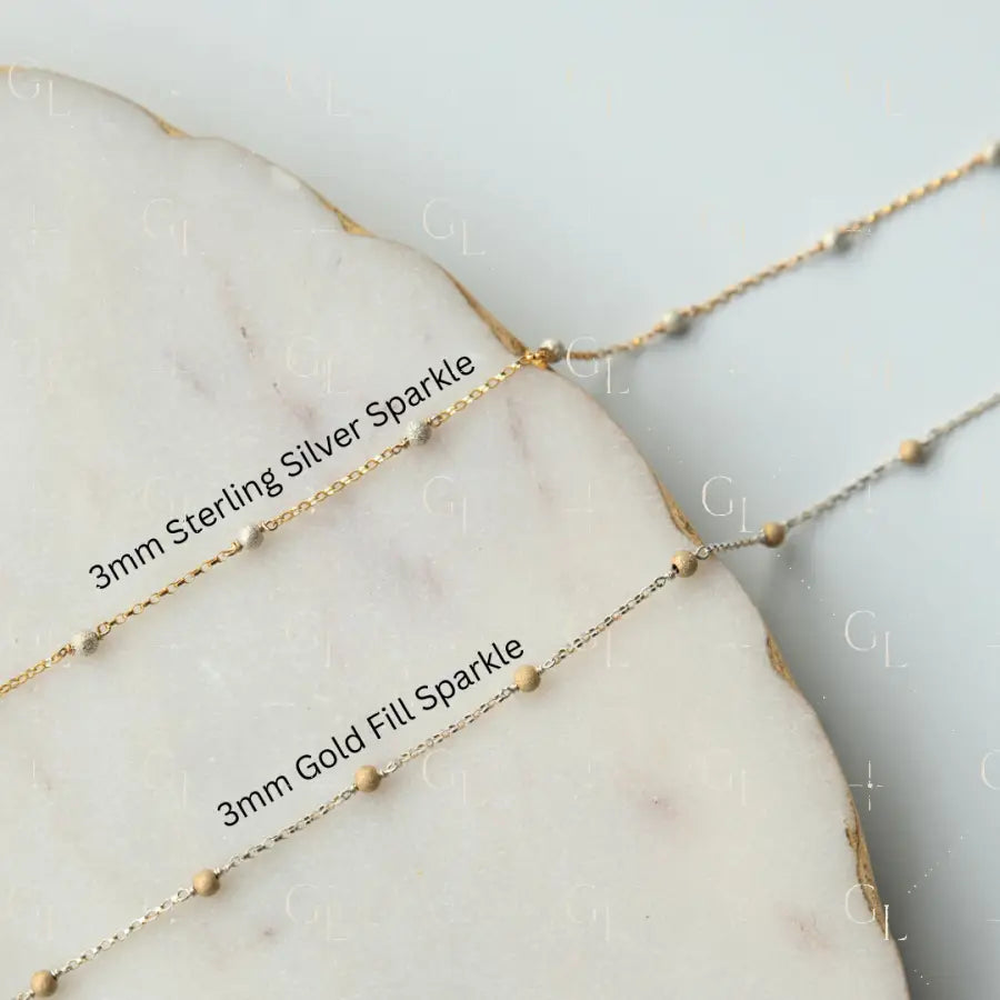 Two gold necklaces with '3mm Sterling Silver Sparkle' and '3mm Gold Fill Sparkle' labels on a marble surface.